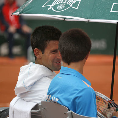 Novak Djokovic sonrie con un recoge pelotas a quien invita a sentarse a su lado mientras escampa